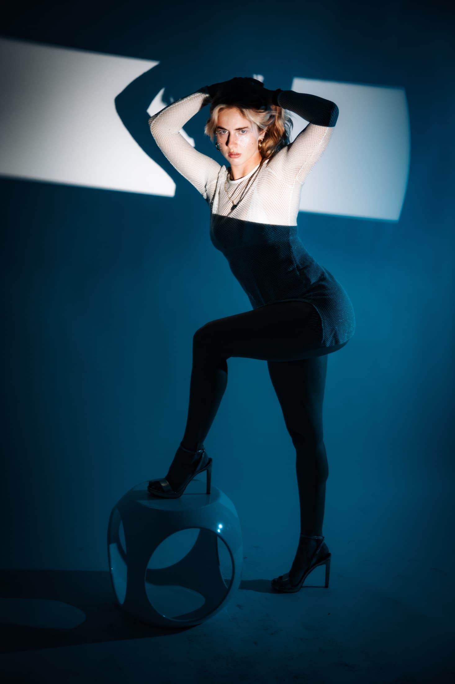 A studio portrait of a woman posed on a blue sphere beneath a hard geometric shadow.