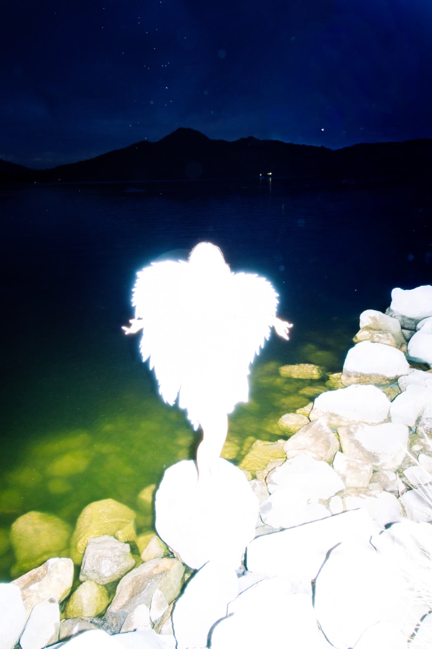 An overexposed figure beside rocks and a dark blue lake at night.